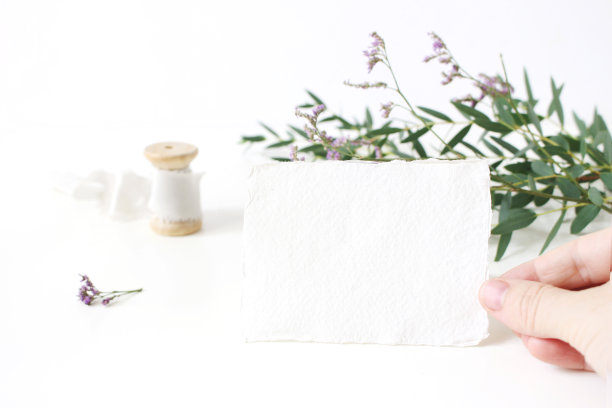 明亮的女性婚礼文具模型场景。女人手握手工纸放置卡的特写。白色的桌子背景上是一束丝带、桉树叶和紫色的补血草花。图片下载