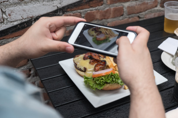 近距离观察，一名男子正在户外酒吧用手机拍摄食物。用手机给食物拍张照片。汉堡、薯条和啤酒放在外面的白色盘子里，放在黑色的桌子上，专注于食物图片下载