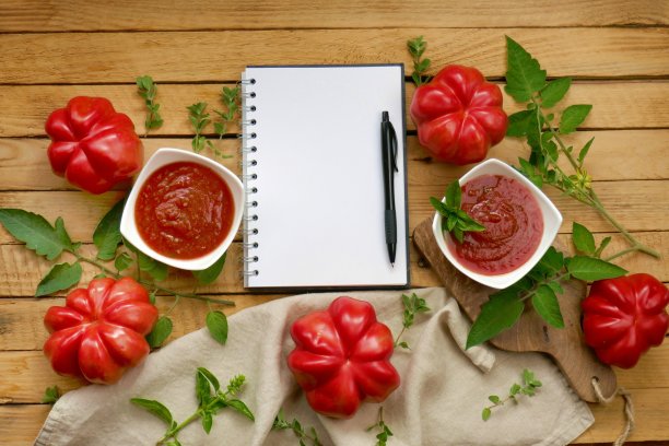 番茄酱。番茄酱。番茄酱的配方。白盘子里装着自制的番茄酱，成熟的红色大西红柿，叶子，罗勒，马角兰，木桌上放着一个空笔记本。图片下载