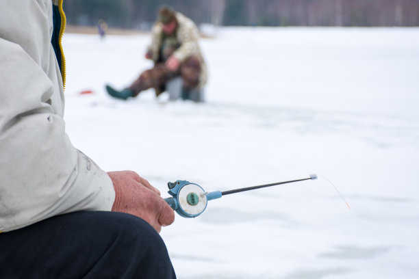 前景中，渔夫手拿钓竿坐着，背景中是渔民在冰上模糊的剪影。这是渔民冬季在冰上钓鱼的照片图片下载
