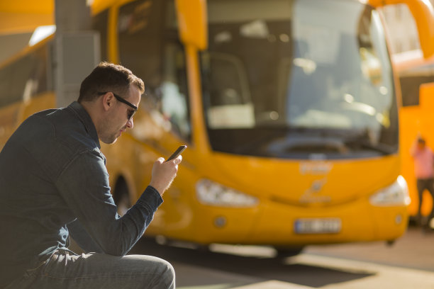 一幅年轻英俊的微笑男子手拿现代设计的智能手机在车站等车的肖像。乘坐环保汽车出行。美好的夏天，阳光明媚的天气和温暖的色彩照片。图片下载