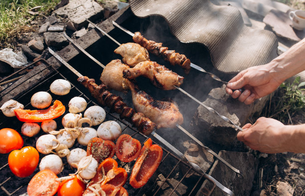 男人手里拿着烤蔬菜、鸡肉和猪肉在明火上。烤架上放洋葱，蘑菇，番茄和红辣椒。砖做的烧烤场所。夏日美食照片图片下载