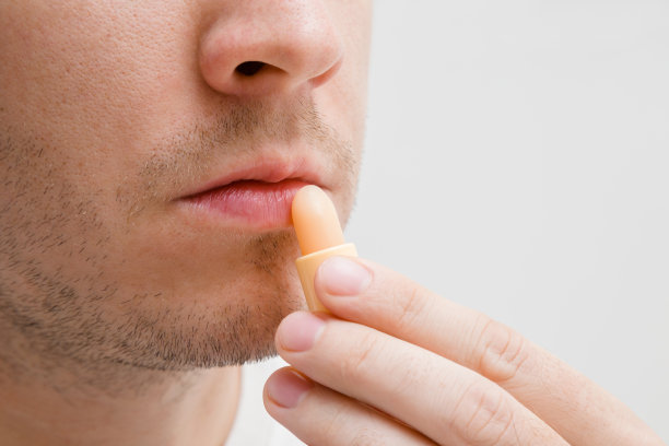 英俊，年轻，蓄胡须的男子使用自然唇膏在浅灰色背景。呵护健康、滋润、柔软的双唇。防止寒冷的冬天，炎热的夏天和多风的秋天。图片下载