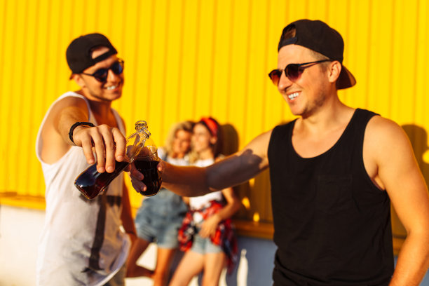 照片背景是一群朋友，一群年轻人拿着玻璃瓶在超市旁边摆着一个饮料摊，在黄色的背景上，朋友们走出来饱满，心情开朗，阳光明媚，生活理念图片下载