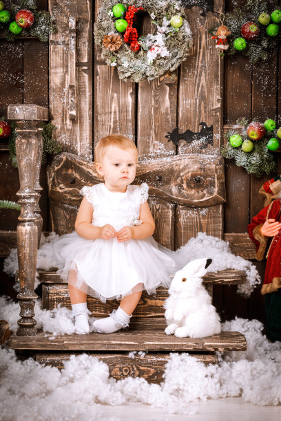 快乐的小女孩穿着白色的连衣裙坐在圣诞节木制的棕色装饰的房间内部图片下载