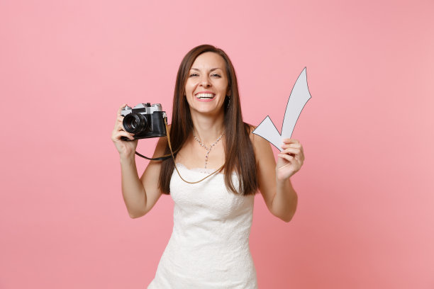 微笑有趣的新娘穿着婚纱拿着复古复古的照片相机和检查标记，选择工作人员，摄影师孤立在粉红色的背景。婚礼待办事项清单。组织的庆祝。图片下载