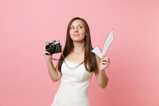 沉思的新娘在婚纱上抬头持有复古复古的照片相机和检查标记选择工作人员摄影师孤立在粉红色的背景。婚礼待办事项清单。组织的庆祝。图片下载