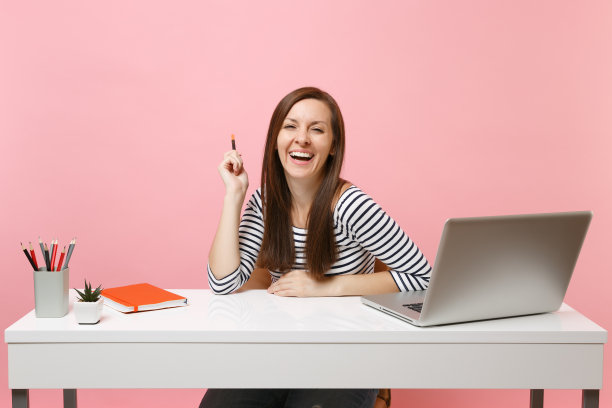 年轻快乐的笑女人，穿着休闲衣服，拿着铅笔，坐在白色的办公桌上工作，当代的电脑笔记本电脑在淡粉色的背景孤立。成就事业理念。副本的空间。图片下载