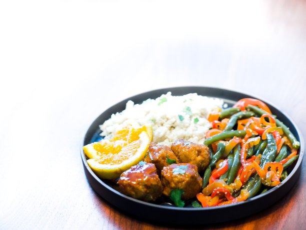 这是一张低碳水化合物肉丸晚餐的特写照片，配以捣碎的花椰菜、菜豆、红辣椒、橙色薄片和香菜，放在一个以木头为背景的黑色盘子里图片下载