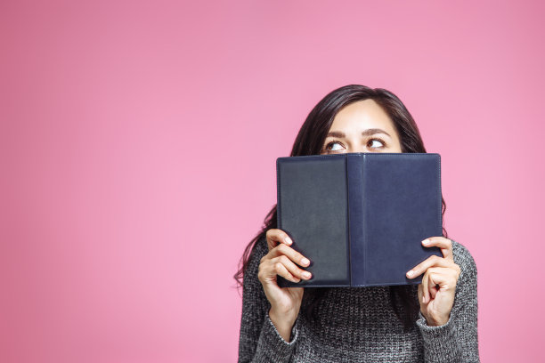 近距离的肖像，一个年轻的女人覆盖她的嘴与书和看远离孤立在粉红色的背景图片下载