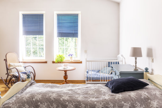 婴儿室内部有一张成人床，一张白色婴儿床和一把摇椅。背景上有百叶窗的窗户。真实的照片。图片下载