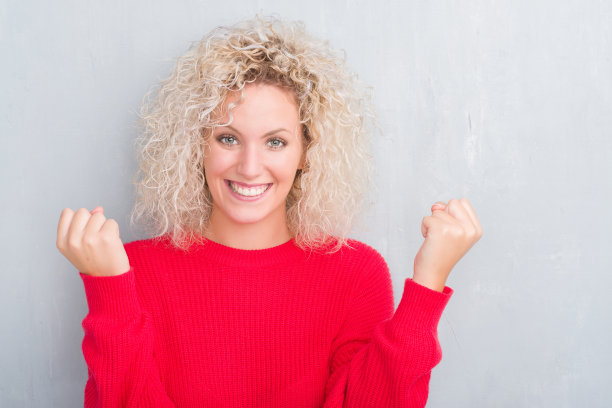 年轻的金发女子与卷发在垃圾灰色背景庆祝惊讶和惊讶的成功，举起手臂和睁开眼睛。赢家的概念。图片下载