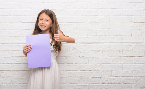 年轻的西班牙裔孩子在白色的砖墙上拿着粉红色的纸，开心地笑着做ok的手势，用手指竖起大拇指，优秀的手势图片下载