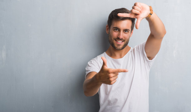 英俊的年轻人越过灰色垃圾墙微笑，用手和手指做框架与快乐的脸。创意和摄影概念。图片下载