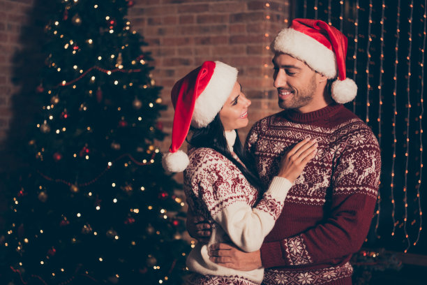 侧视图照片的黑发女士，英俊男子在装饰毛衣站在客厅舒适，舒适的房子内部与灯光花环装饰松树三互相看和拥抱图片下载