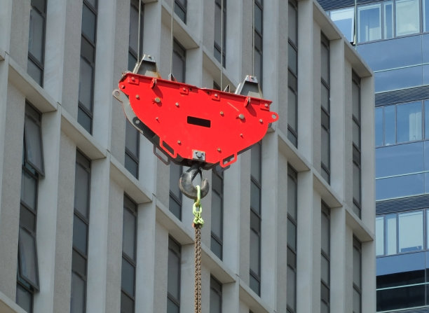在一个以城市现代建筑为背景的建筑工地上，一个红色的吊钩和滑轮正在吊起一个悬挂的金属链图片下载