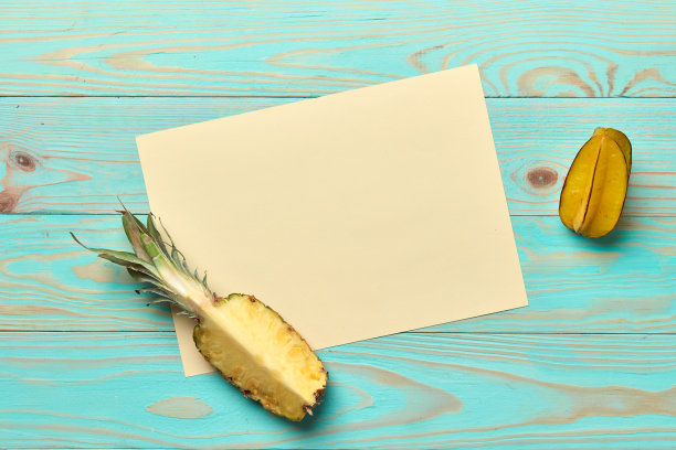 白色的硬纸板上有一片菠萝和一整个杨桃，背景是蓝色的木头，有文字空间。平躺图片下载