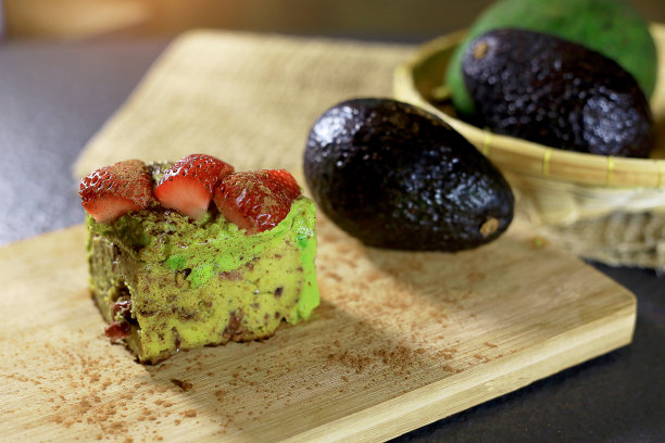 鳄梨蛋糕的特写，上面有草莓，杏仁粉，蔓越莓，蓝莓，里面。木桌上放着肉桂粉。还有篮子里的牛油果。健康或生酮甜点概念。黑暗的基调。图片下载