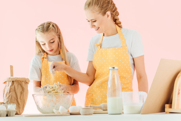 微笑的母亲和可爱的小女儿在黄色围裙准备面团一起孤立在粉红色图片下载