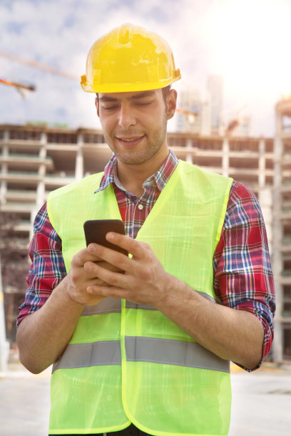 年轻工程师在建筑工地和起重机背景下使用智能手机。图片下载