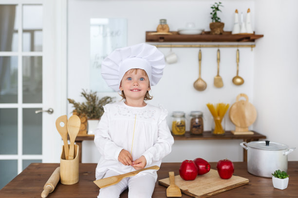 戴着厨师帽的小女孩坐在摆满红苹果的厨房桌子上。健康食品和健康生活方式的概念。图片下载