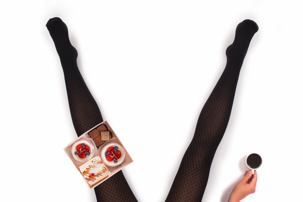 创意照片的女性腿在黑色连裤袜与一杯咖啡和一盒小蛋糕在白色的背景。的概念。图片下载