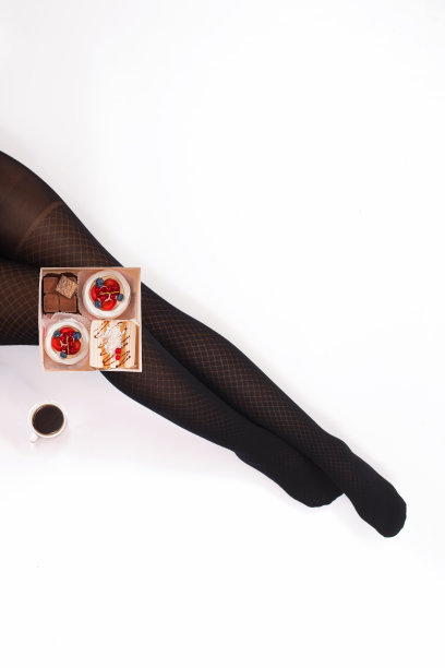 创意照片的女性腿在黑色连裤袜与一杯咖啡和一盒小蛋糕在白色的背景。的概念。图片下载