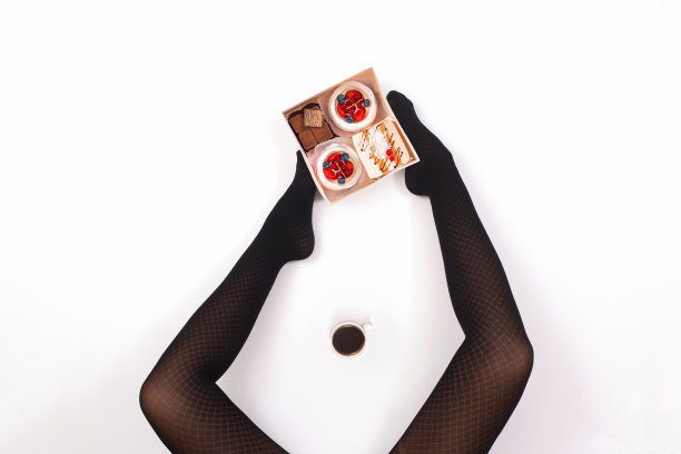 创意照片的女性腿在黑色连裤袜与一杯咖啡和一盒小蛋糕在白色的背景。的概念。图片下载