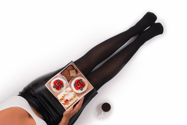 创意照片的女性腿在黑色连裤袜与一杯咖啡和一盒小蛋糕在白色的背景。的概念。图片下载