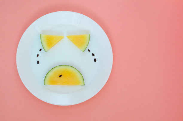 哭泣的脸，眼睛和嘴巴是用黄色的西瓜和种子上的泪水放在一个白色的盘子和粉红色的背景。图片下载