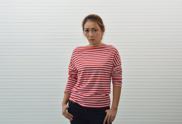 穆迪年轻的亚洲女人穿着红白条纹衬衫和牛仔裤站在白色墙壁的背景，不安的面部表情图片下载