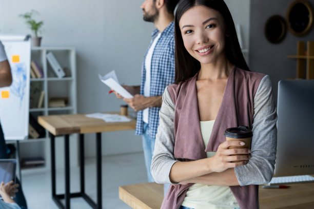 自信而成功的领导者。自信的年轻女子双臂交叉，微笑着看着镜头，她的同事在后面工作。图片下载