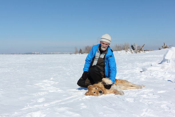 一个留着胡子、穿着蓝色夹克的老人和一只拉布拉多犬在冬天雪地上图片下载