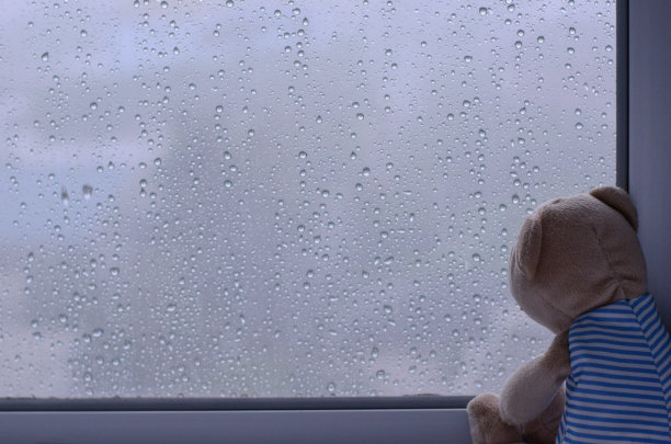 一只悲伤的泰迪熊独自坐在窗前，雨水打在玻璃上。照片有文字空间。图片下载