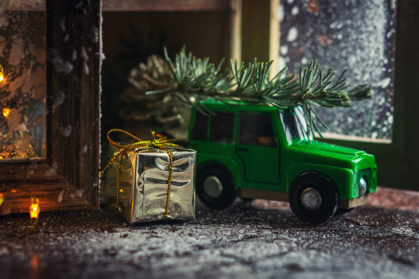 金色的礼盒和圣诞装饰品，绿色的玩具车载着一棵圣诞树，木质桌子上有白色的背景。圣诞节庆祝和新年的概念。图片下载