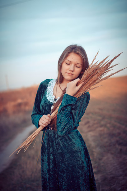 一个长头发的女孩，穿着绿色的裙子，手里拿着一束干草枝。站在田野附近的一条乡间小路上。时间是秋天。图片下载