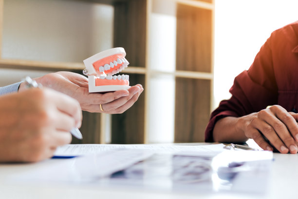 一个自信的牙医拿着假牙，在办公室里和老病人交谈。图片下载