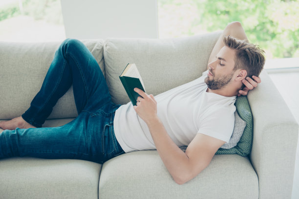 侧面的照片有吸引力，留胡子，好看的男人在白t恤躺在床上，读书人辛苦工作一周后在轻现代室内图片下载