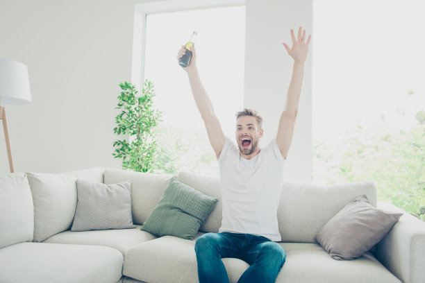 有吸引力的照片，运动的男人在牛仔裤和白色t恤坐在沙发在轻现代室内看电视举起他的手图片下载