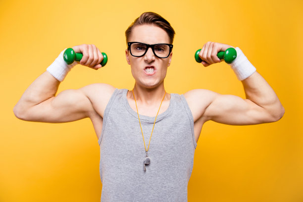 我能做到!一个年轻男子的特写照片被孤立在生动的黄色背景上，他咬紧牙关，用绿色的低重量哑铃推着双手。图片下载