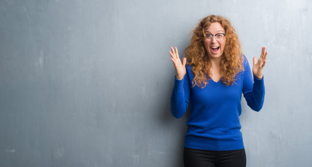 年轻的红头发的女人越过灰色的垃圾摇滚墙庆祝疯狂和对成功的惊讶，举起手臂，睁开眼睛兴奋地尖叫。赢家的概念图片下载