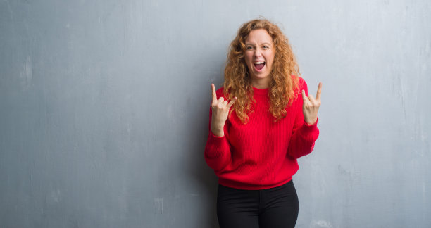 年轻的红发女人越过灰色垃圾墙穿着红色毛衣大喊与疯狂的表情做摇滚象征与手向上。音乐明星。沉重的概念。图片下载