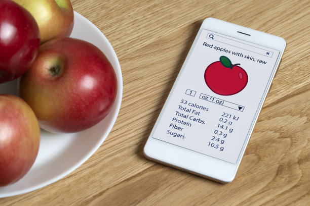 白色智能手机，屏幕上有一款计算卡路里的应用程序，屏幕位于放苹果的盘子附近的一个木头表面上。图片下载