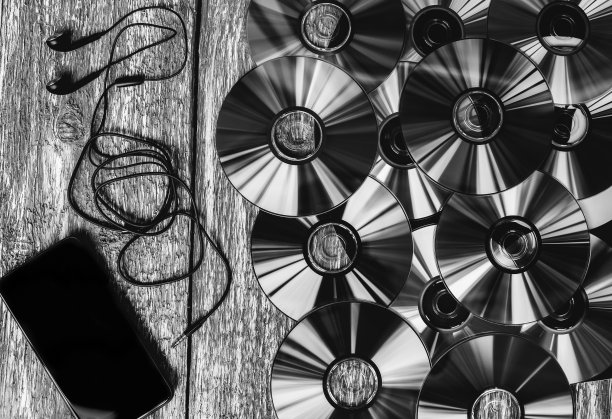 许多Cd光盘躺在古老的木头桌子上。全息颜色。黑色的智能手机和耳机就在附近。音乐和现代科技照片。数字的动机。黑色和白色。图片下载