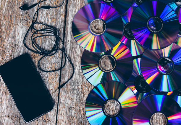 许多Cd光盘躺在古老的木头桌子上。全息颜色。黑色的智能手机和耳机就在附近。音乐和现代科技照片。数字的动机。图片下载