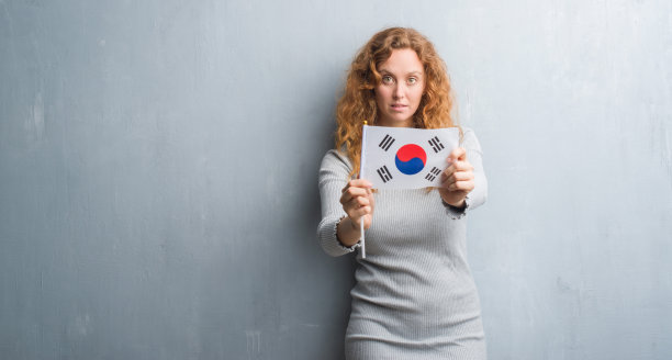 年轻的红发女子在灰色墙举着韩国国旗自信的表情在聪明的脸上思考严肃图片下载