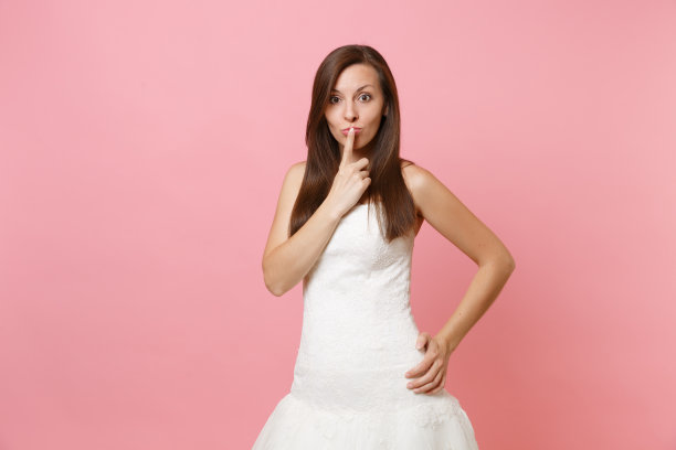 美丽的新娘的肖像在白色蕾丝婚纱站和保持食指在嘴唇孤立的粉色柔和的背景。婚礼庆典的概念。广告版面复制。图片下载
