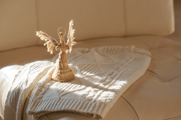 俯视图图像的白色舒适针织毛衣与天使在阳光下米色皮革沙发背景。舒适的圣诞毛衫。温暖舒适的冬天衣服。圣诞礼物的想法。副本的空间。图片下载