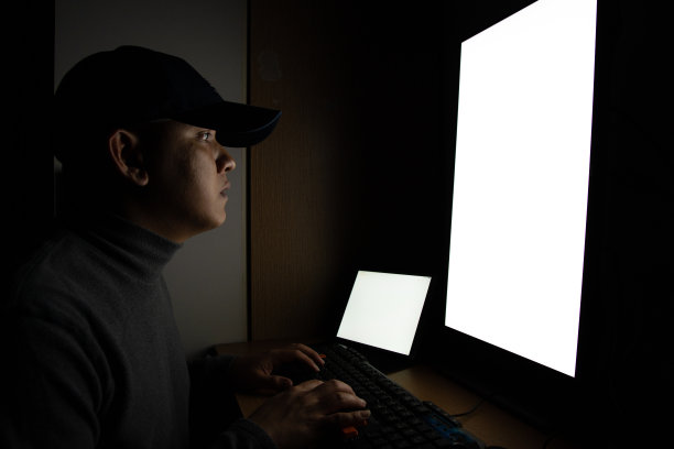 侧视图男子黑客坐在电脑显示器和白色屏幕平板电脑在黑暗的房间。图片下载
