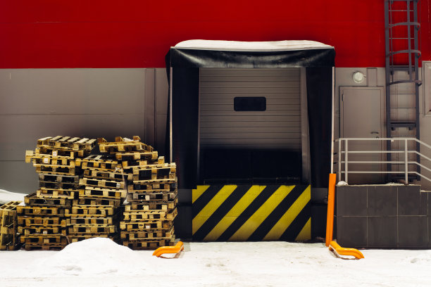 有黄黑对角线警告标志的储货升降机。雪地上的木托盘。有拷贝空间的自动装货门。冬天仓库的红墙。近距离的工业建筑图片下载